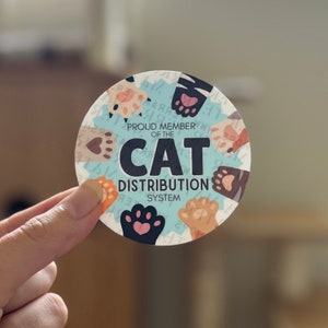 May include: A round sticker with a blue background and a white border. It features a paw print design with the text "Proud Member of the CAT Distribution System".