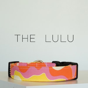 May include: A colorful dog collar with a pink, orange, and yellow abstract pattern. The collar has a black buckle and a gold metal ring. The text "THE LULU" is printed above the collar.