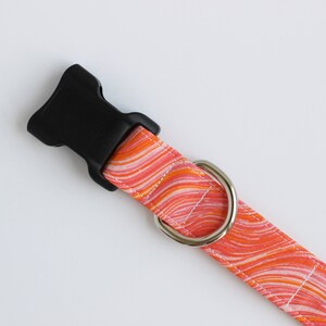 May include: A pet collar featuring a vibrant orange, pink, and white marbled wave pattern. The collar includes a black plastic side-release buckle and a polished silver-toned metal D-ring for attachment, secured with white stitching along the edges.