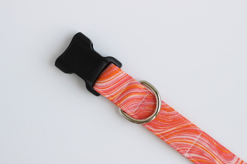 May include: A pet collar featuring a vibrant orange, pink, and white marbled wave pattern. The collar includes a black plastic side-release buckle and a polished silver-toned metal D-ring for attachment, secured with white stitching along the edges.