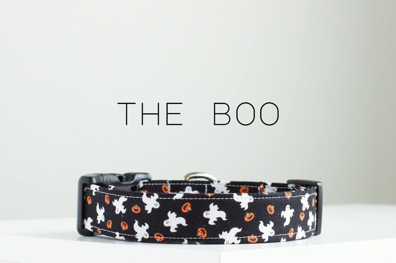 May include: Black dog collar with a Halloween themed print featuring white ghosts and orange pumpkins. The collar has a black buckle and the text "THE BOO" is printed above the collar.