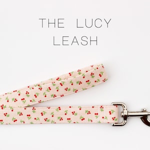 May include: The Lucy Leash. A light pink dog leash with a cherry pattern in red and green. The leash has a silver metal clasp. The text "THE LUCY LEASH" is at the top of the image.