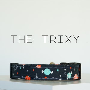 Puede incluir: Un collar de perro negro con un patrón colorido de caramelos, estrellas y calabazas. El texto "THE TRIXY" está impreso sobre el collar.