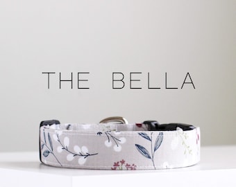 May include: A beige dog collar with a floral pattern in white, blue, and red. The collar has a silver buckle and black adjustable strap. The text "THE BELLA" is printed above the collar.