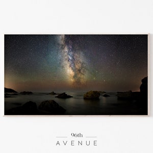 Może przedstawiać: Zdjęcie Drogi Mlecznej nad ciemnym, spokojnym zbiornikiem wodnym. Woda jest spokojna i odbija gwiazdy na niebie. W wodzie znajduje się kilka ciemnych skał, a linia brzegowa jest widoczna w oddali.