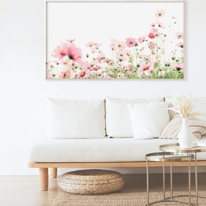 May include: A framed print of a field of pink cosmos flowers against a white background. The print is hanging on a white wall above a white couch with pillows and a wooden coffee table.