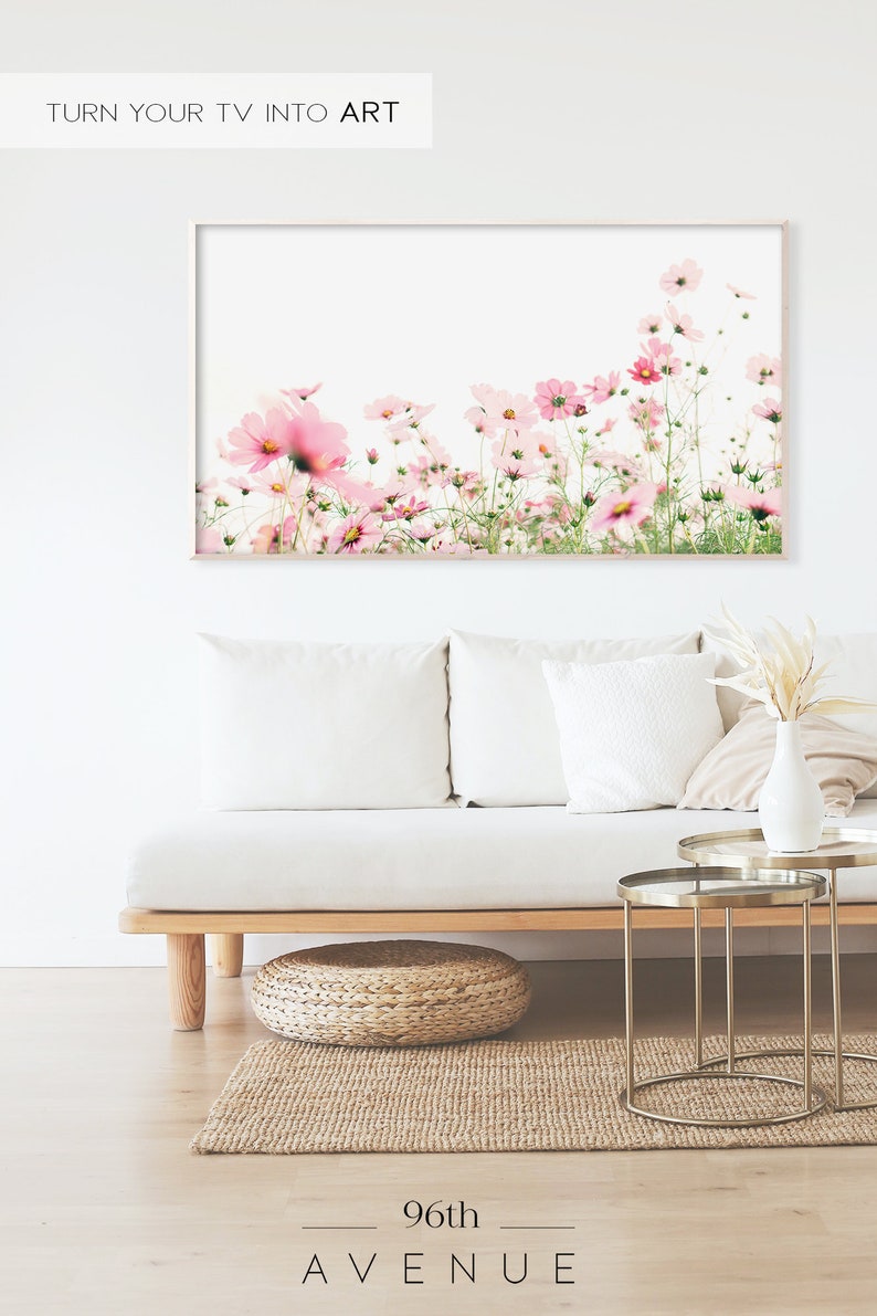 May include: A framed print of a field of pink cosmos flowers against a white background. The print is hanging on a white wall above a white couch with pillows and a wooden coffee table.