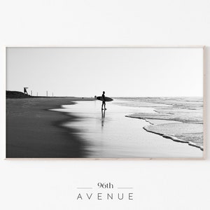 May include: Black and white photograph of a surfer walking on a beach with a surfboard under their arm. The ocean is in the background with waves crashing on the shore. The photo is framed in a light wood frame.