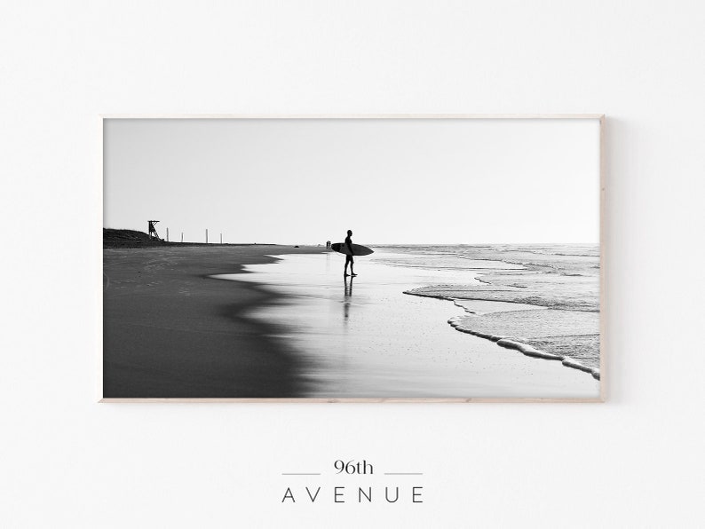 May include: Black and white photograph of a surfer walking on a beach with a surfboard under their arm. The ocean is in the background with waves crashing on the shore. The photo is framed in a light wood frame.