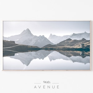 Puede incluir: Una fotografía enmarcada de una cordillera reflejada en un lago tranquilo. Las montañas están cubiertas de nieve y el lago es de color azul claro. La imagen está enmarcada en un marco de madera claro.