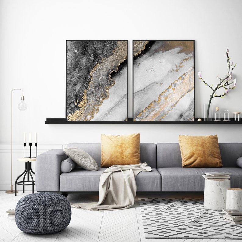 Puede incluir: Dos impresiones de arte abstracto en marcos negros, con un fondo blanco y gris con acentos dorados. Las impresiones se muestran en un estante sobre un sof&aacute; gris con almohadas y una manta.