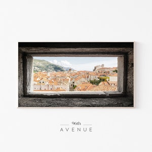 Puede incluir: Una vista de una ciudad a través de una ventana de piedra. La ciudad está formada por muchos edificios con techos de tejas rojas. La ventana está enmarcada en piedra y tiene una textura rugosa.