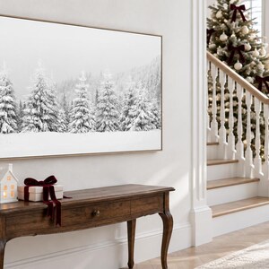 K&ouml;nnte beinhalten: Ein rechteckiger Wandkunstdruck mit einer schwarz-wei&szlig;en Winterlandschaft aus schneebedeckten Tannenb&auml;umen. Der Druck ist in einem hellen Holzrahmen gefasst und an einer wei&szlig;en Wand &uuml;ber einem dunklen Holzkonsolentisch angebracht.