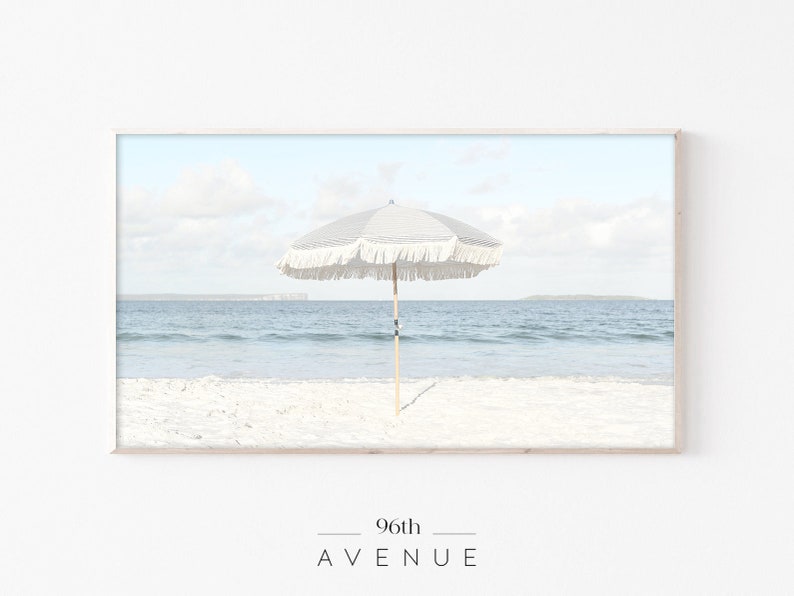 May include: A white beach umbrella with fringe trim stands on a white sandy beach with blue ocean water in the background.