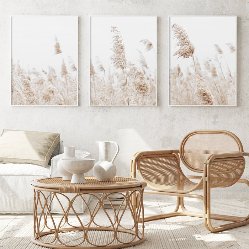 May include: Three framed prints of beige dried grass blowing in the wind. The prints are hanging on a white wall above a wicker chair and a round wicker coffee table.
