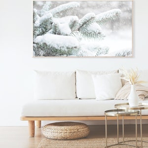 May include: A snowy winter scene with pine branches covered in white snow. The branches are in focus, while the background is blurred and out of focus. The image is framed in a light wood frame.