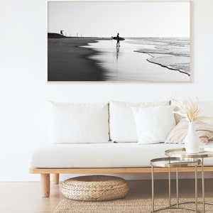 May include: Black and white photograph of a surfer walking on a beach with a surfboard under their arm. The photo is framed and hanging on a white wall above a white couch.