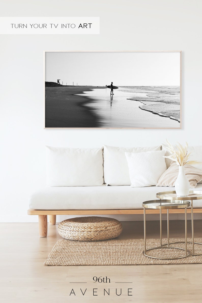 May include: Black and white photograph of a surfer walking on a beach with a surfboard under their arm. The photo is framed and hanging on a white wall above a white couch.