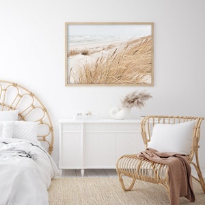 May include: A white bedroom with a wicker headboard bed, a white dresser, a wicker chair with a brown throw blanket, and a framed photograph of a beach scene with tall grass.