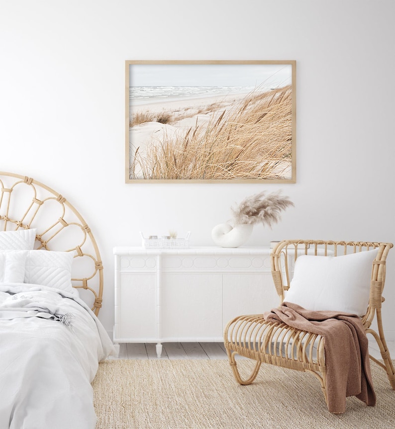 May include: A white bedroom with a wicker headboard bed, a white dresser, a wicker chair with a brown throw blanket, and a framed photograph of a beach scene with tall grass.
