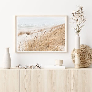 May include: A framed photograph of a beach scene with tall grasses in the foreground and a blue ocean in the background. The photo is displayed on a light wood shelf with a white vase, a small wooden bowl, and a white cotton stem.