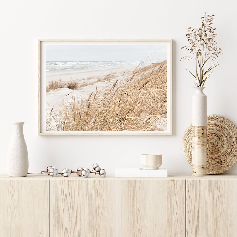 May include: A framed photograph of a beach scene with tall grasses in the foreground and a blue ocean in the background. The photo is displayed on a light wood shelf with a white vase, a small wooden bowl, and a white cotton stem.