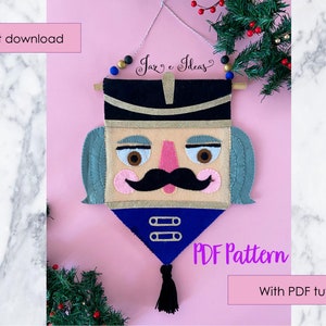 Pode incluir: Um pendurador de parede de feltro com um quebra-nozes, com chapéu e bigode pretos, detalhes dourados e detalhes azuis. O texto "PDF Pattern" e "Instant download" são visíveis. O pendurador é decorado com uma borla preta e contas.