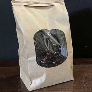 May include: A brown paper bag with a clear window, filled with dark soil. The bag is sealed at the top and stands upright on a wooden surface. The window reveals the contents within.