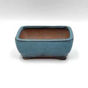 May include: A square blue ceramic bonsai pot with a brown interior and a slightly raised rim. The pot has three small feet on the bottom.