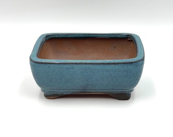 May include: A square blue ceramic bonsai pot with a brown interior and a slightly raised rim. The pot has three small feet on the bottom.