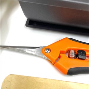 May include: Orange handled pruning shears with a stainless steel blade. The shears have a spring mechanism and a black lock.