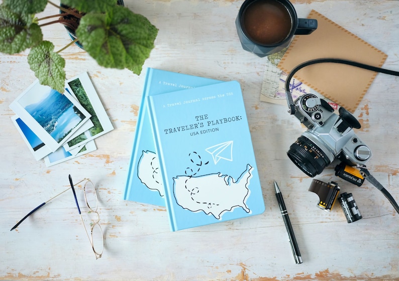 Puede incluir: Un diario de viaje azul con el t&iacute;tulo "The Traveler's Playbook: USA Edition" y un mapa de los Estados Unidos con un avi&oacute;n de papel en la parte superior. El diario est&aacute; sobre una mesa de madera blanca con una c&aacute;mara, cartuchos de pel&iacute;cula, una taza de caf&eacute; y una planta.