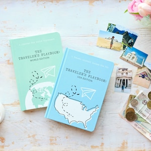 Puede incluir: Dos diarios de viaje azules y verdes con el t&iacute;tulo "The Traveler's Playbook" sobre una superficie de madera blanca. El diario de la edici&oacute;n mundial es verde y presenta un mapa del mundo con un avi&oacute;n de papel. El diario de la edici&oacute;n de EE. UU. es azul y presenta un mapa de los Estados Unidos con un avi&oacute;n de papel.