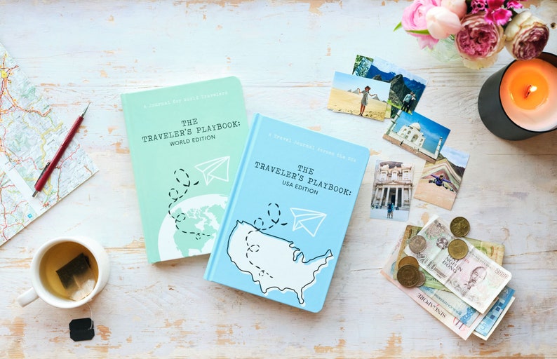 Puede incluir: Dos diarios de viaje azules y verdes con el t&iacute;tulo "The Traveler's Playbook" sobre una superficie de madera blanca. El diario de la edici&oacute;n mundial es verde y presenta un mapa del mundo con un avi&oacute;n de papel. El diario de la edici&oacute;n de EE. UU. es azul y presenta un mapa de los Estados Unidos con un avi&oacute;n de papel.
