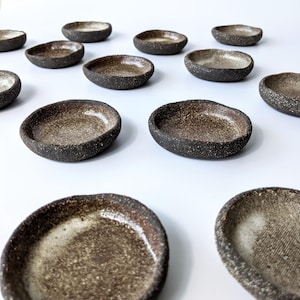 May include: A selection of small, handmade ceramic dishes. The oval-shaped dishes have a speckled, dark brown exterior and a lighter brown interior. The edges are slightly raised, giving a rustic and earthy appearance.