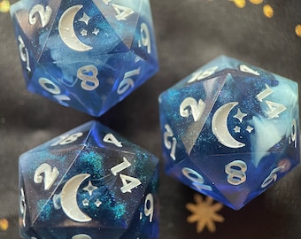 May include: Three blue and white dice with a crescent moon and stars design. The dice are made of a translucent material and have a glittery finish.