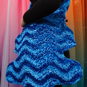 以下が含まれることがあります： ユニークな抽象的な形をした青いベルベットのベスト。ベストは、質感のある波状のパターンと黒いストラップが付いています。