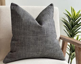 May include: A square throw pillow featuring a dark charcoal gray and black herringbone pattern. The fabric has a visible woven texture with a consistent zigzag design across the entire surface of the pillow, which is plumped to create a central crease.