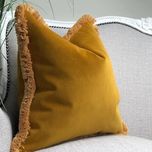 May include: A square mustard yellow velvet throw pillow featuring a matching mustard yellow fringe trim along the edges. The pillow has a smooth, soft-textured surface and is displayed in a upright position, showcasing its vibrant color and detail.