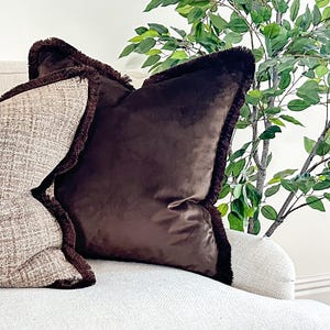 May include: A square throw pillow in a solid dark brown velvet fabric with a dark brown fringe trim along the edges, positioned next to a second throw pillow featuring a textured beige and brown woven pattern with matching fringe trim.