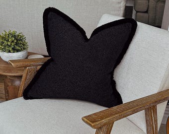 May include: A black throw pillow with a distinct butterfly shape and a textured, fuzzy surface. The pillow features a dark black fringe trim along all of its edges, creating a soft and tactile appearance against the light-colored chair surface.