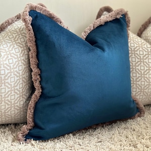 May include: A square throw pillow in deep teal velvet featuring a textured beige fringe trim along all four edges, positioned in front of two other square throw pillows with a beige and white geometric pattern and matching fringe trim.