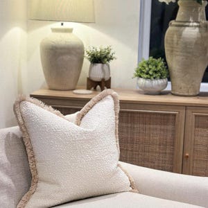 May include: A cream-coloured decorative cushion with a textured surface and a fringed border. The cushion is placed on a light-coloured sofa. A lamp, vases, and plants are visible in the background, suggesting a home decor theme.