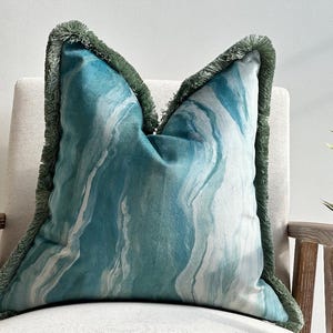 May include: A decorative pillow with a blue and white watercolor design. The pillow has a green fringe trim and is placed on a neutral-colored chair. The pillow's design resembles flowing water.
