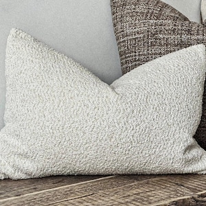 May include: A light-coloured, textured pillow with a soft, plush appearance rests on a wooden surface. Another pillow with a brown, woven pattern is partially visible behind it. The pillows are decorative and suitable for home decor.