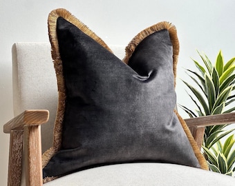 May include: A square throw pillow featuring a dark charcoal gray velvet fabric. The edges of the pillow are finished with a thick, textured golden-brown fringe trim that runs along the perimeter, providing a distinct contrast to the dark surface.