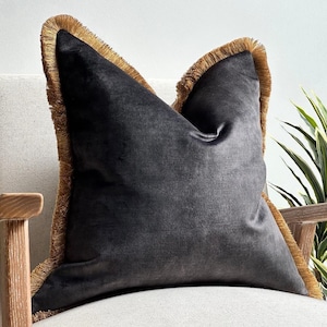 May include: A dark grey velvet cushion with a gold fringe trim. The square cushion is on a cream-coloured chair with wooden arms. A decorative home accent.