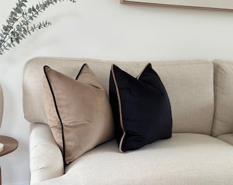 May include: Two square velvet throw pillows. One pillow is a light tan color with black piping along the edges. The second pillow is a solid black color with light tan piping along the edges. Both pillows have a soft, smooth texture.
