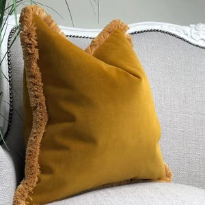May include: Mustard yellow velvet cushion with fringed edges. The square cushion is placed on a light grey upholstered chair. The fringe is a lighter shade of yellow. The cushion adds a pop of colour to the neutral background.