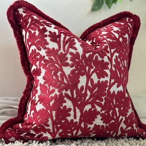 May include: A square throw cushion featuring a cream-coloured base with a dense, raised red velvet floral pattern. The edges of the cushion are finished with a matching red velvet fringe trim, creating a textured border around the entire square shape.